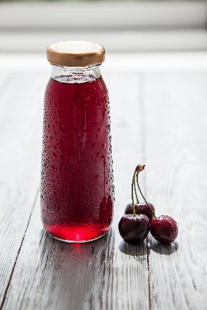 A glass bottle sitting on a table with cherry juice in it and cherries sitting nearby.