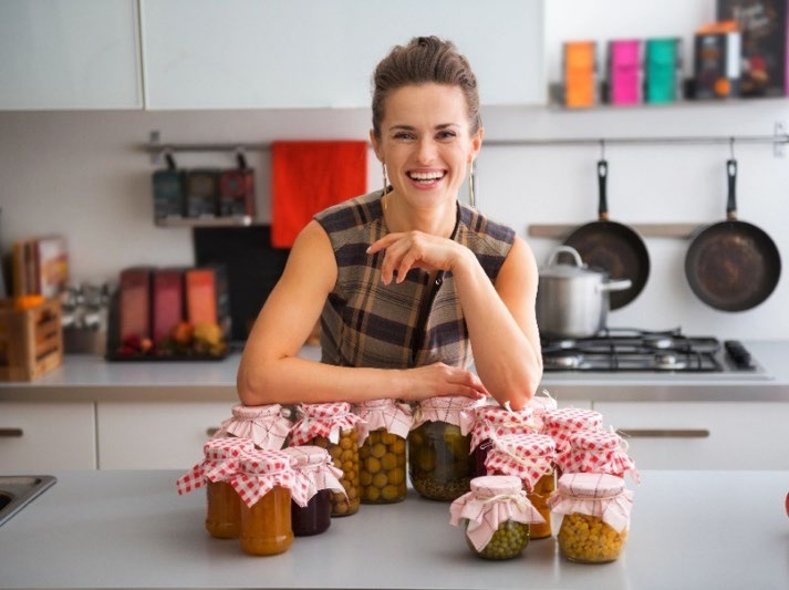 A smiling women in a kitchen standing over a counter with bottled food.