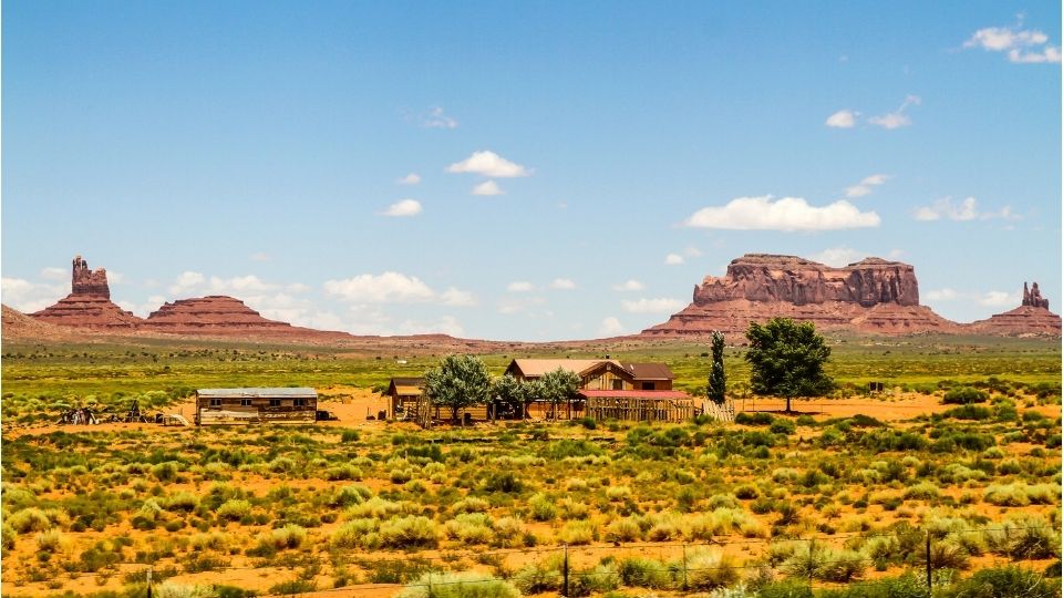 Wide desert landscape with red rock mesas, scattered shrubs, and a small group of buildings under a blue sky.