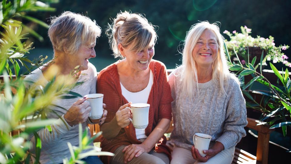 3 middle aged women sitting on an outdoor bench holding teacups.