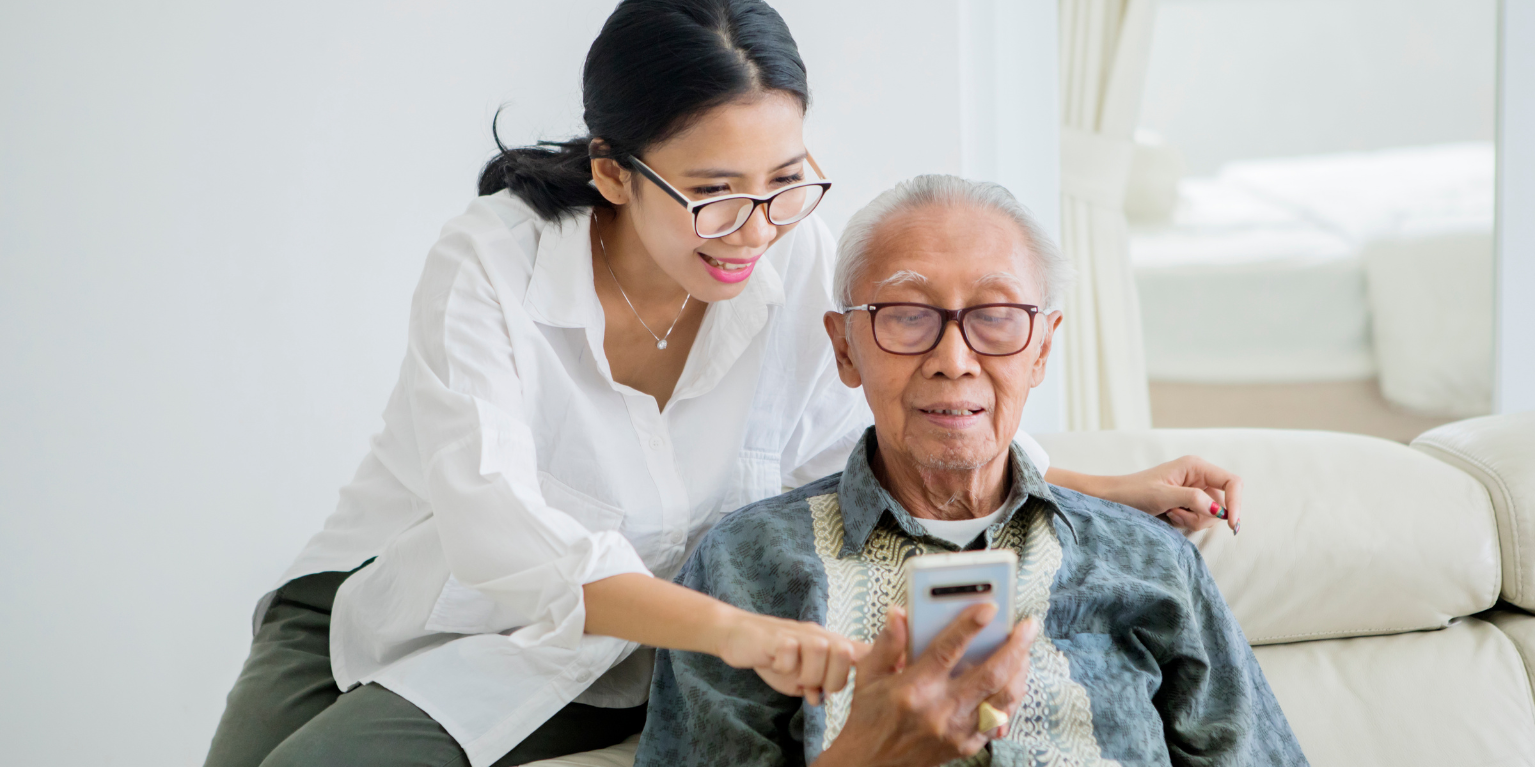 a younger woman assists an elderly man with using his phone while sitting on a white couch 