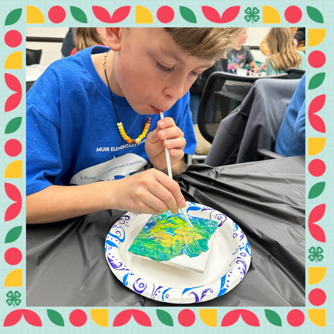 A boy leans over a canvas covered in paint using a straw to blow the paint making a design
