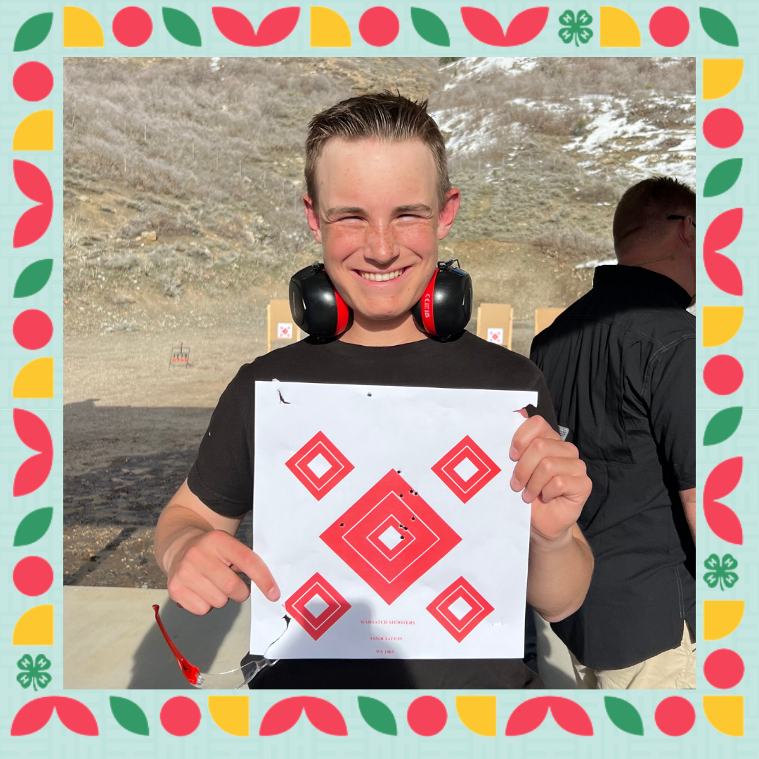 A boy stands in front of a shooting range holding a paper with holes in the center diamond, showing off his aim.