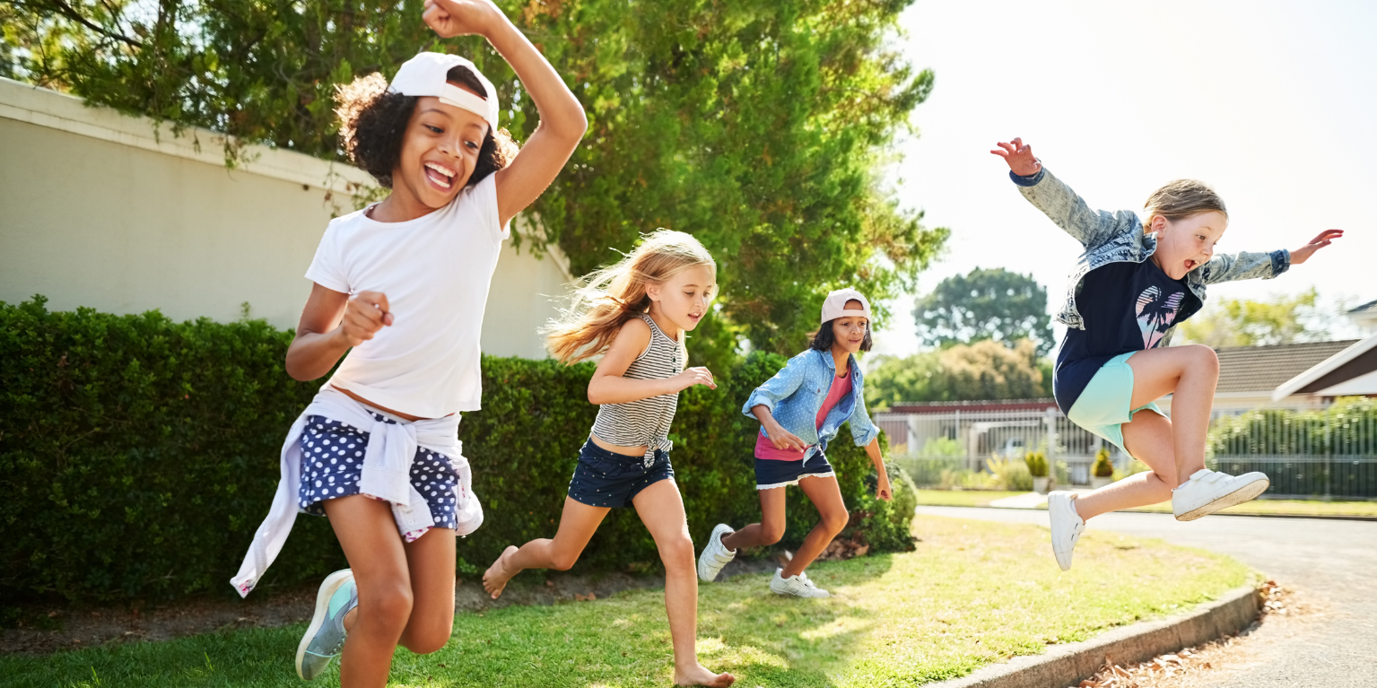 Children running and jumping together on a sunny neighborhood lawn, smiling and playing outdoors near trees and homes on a bright summer day.