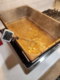 A wide pan of light gold liquid simmers on a stove with a digital thermometer connected to the side