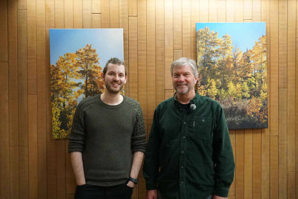 Drew Holley and Darren McAvoy stand in front of a wooden wall with pictures