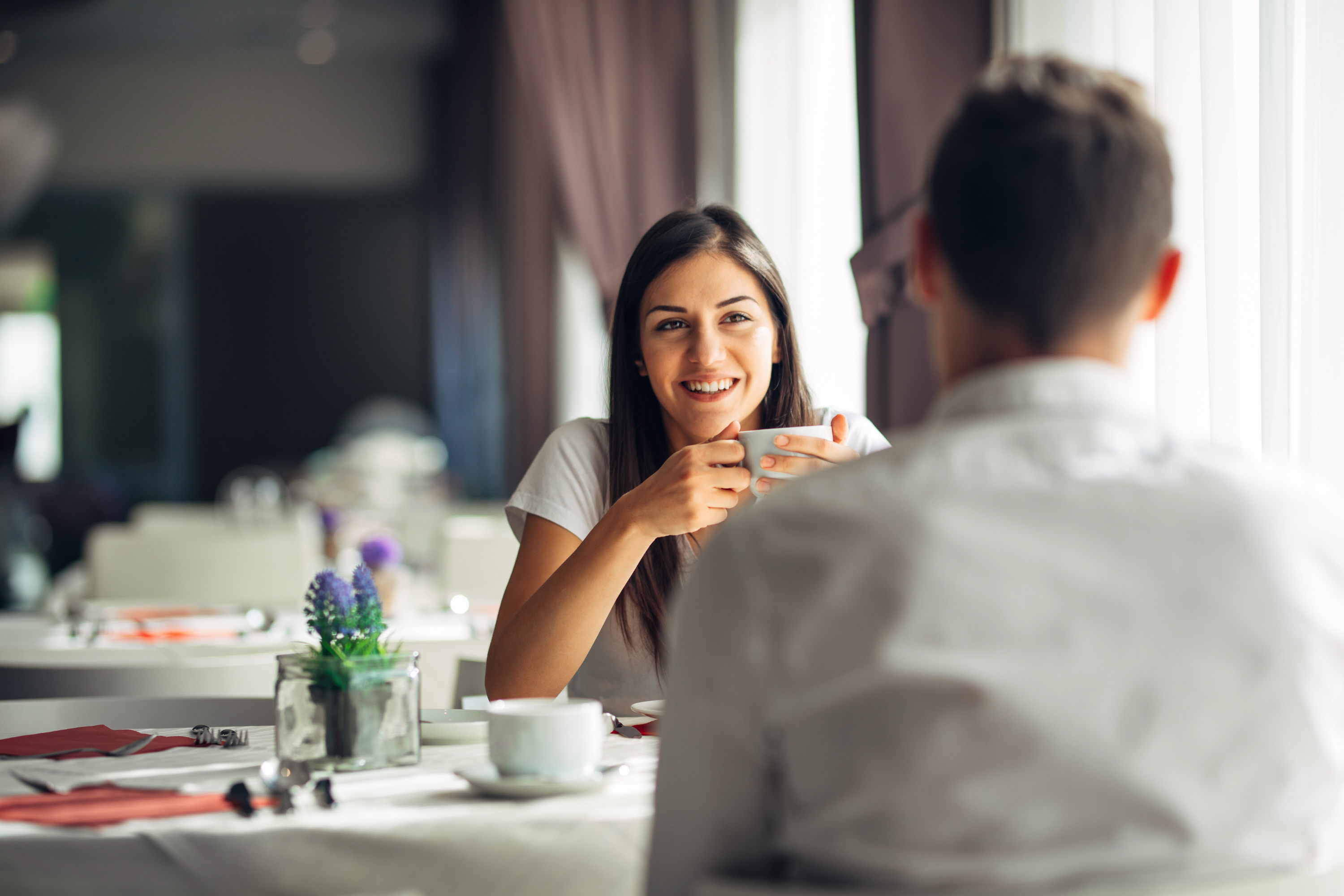 woman smiling while sitting with a coffee cup in her hand facing a man with his back to us on a first date
