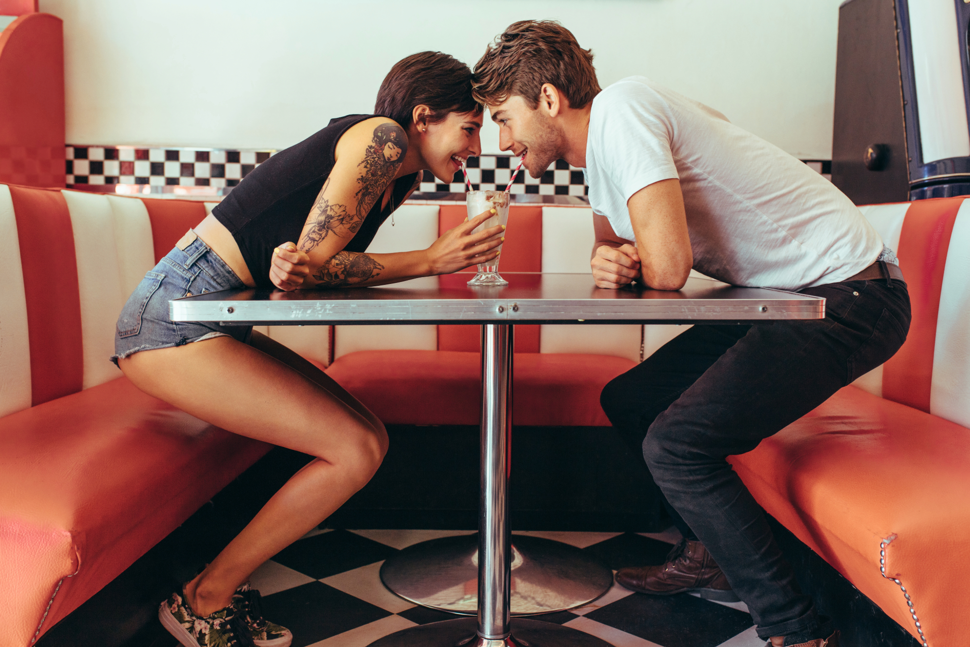 man and woman in a restaurant looking at each other while drinking a milkshake together