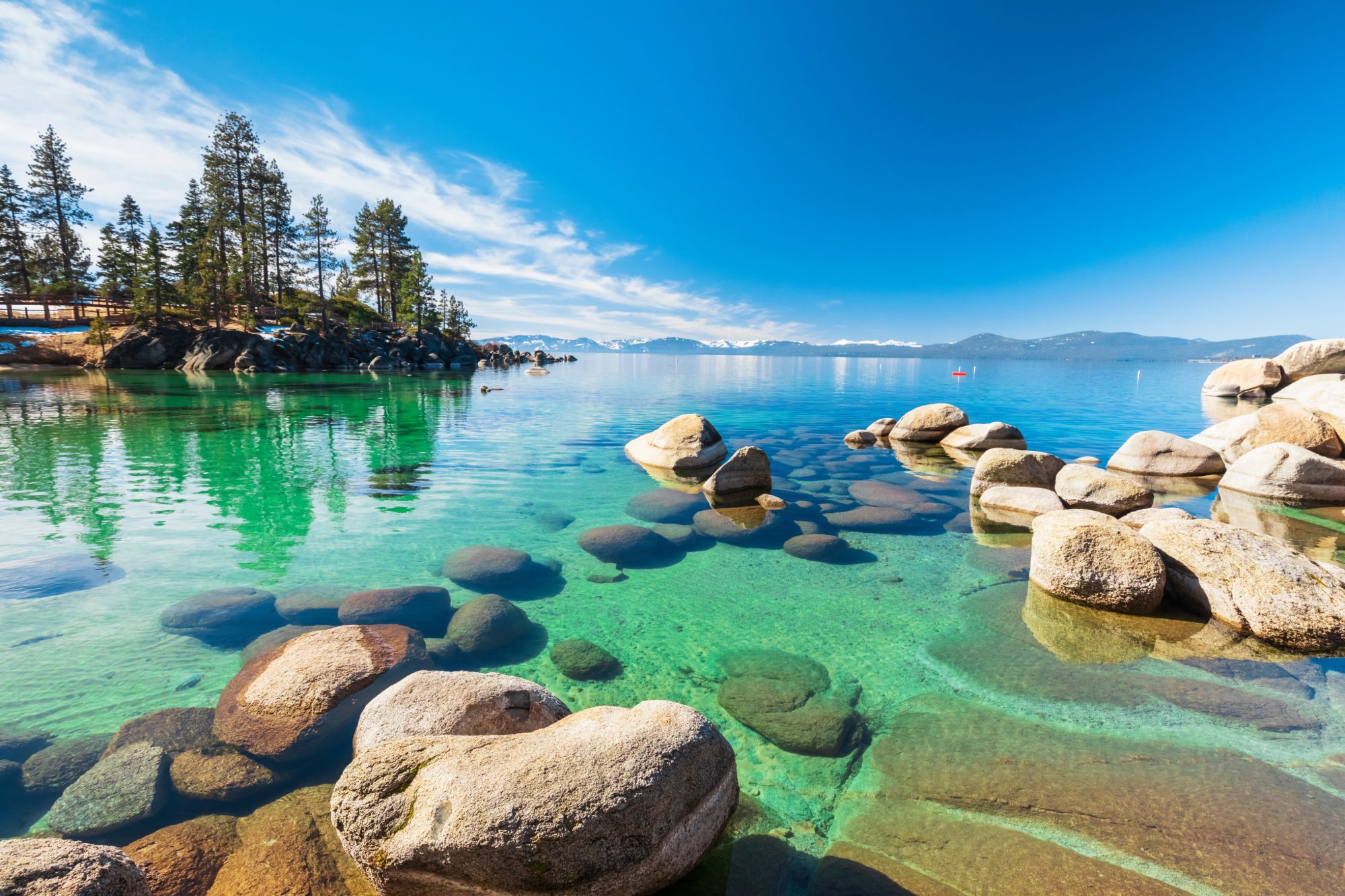 Shoreline of Lake Tahoe featuring crystal clea water, rocks, trees, and mountains far in the background