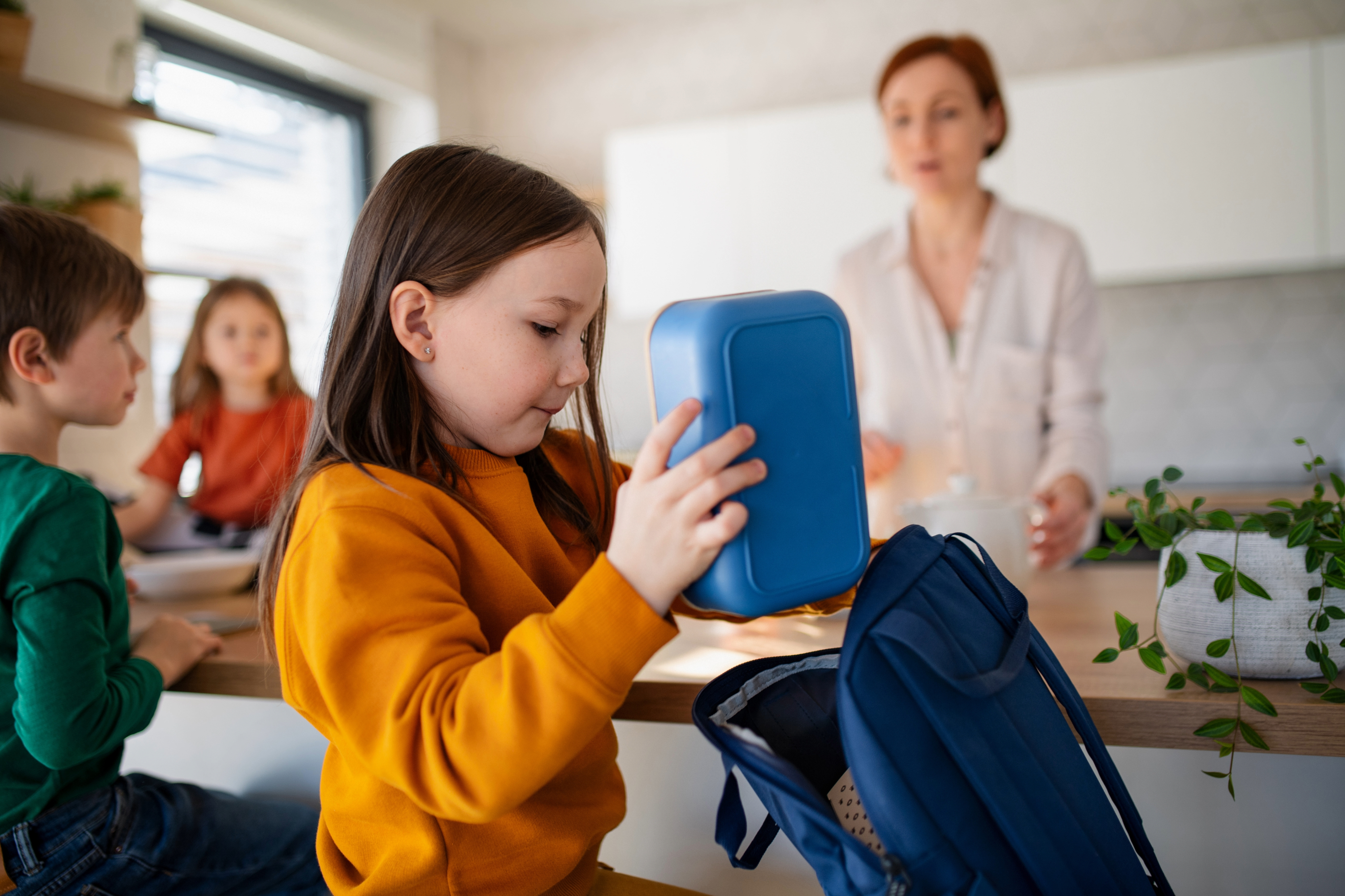 little girl putting her own lunchbox in her backpack under the watchful eyes of her mom