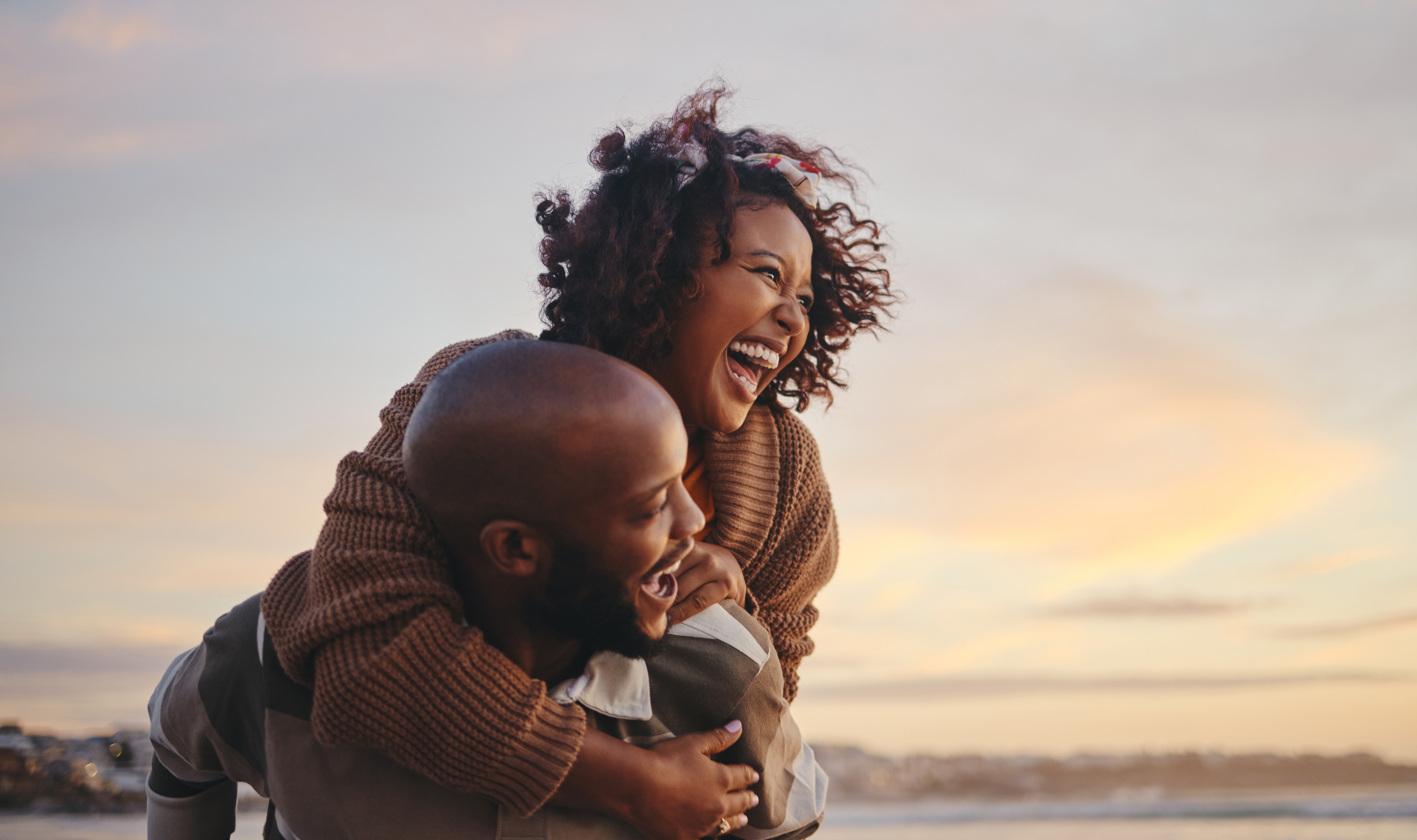 man carrying a woman piggyback-style on the beach during sunset, both laughing