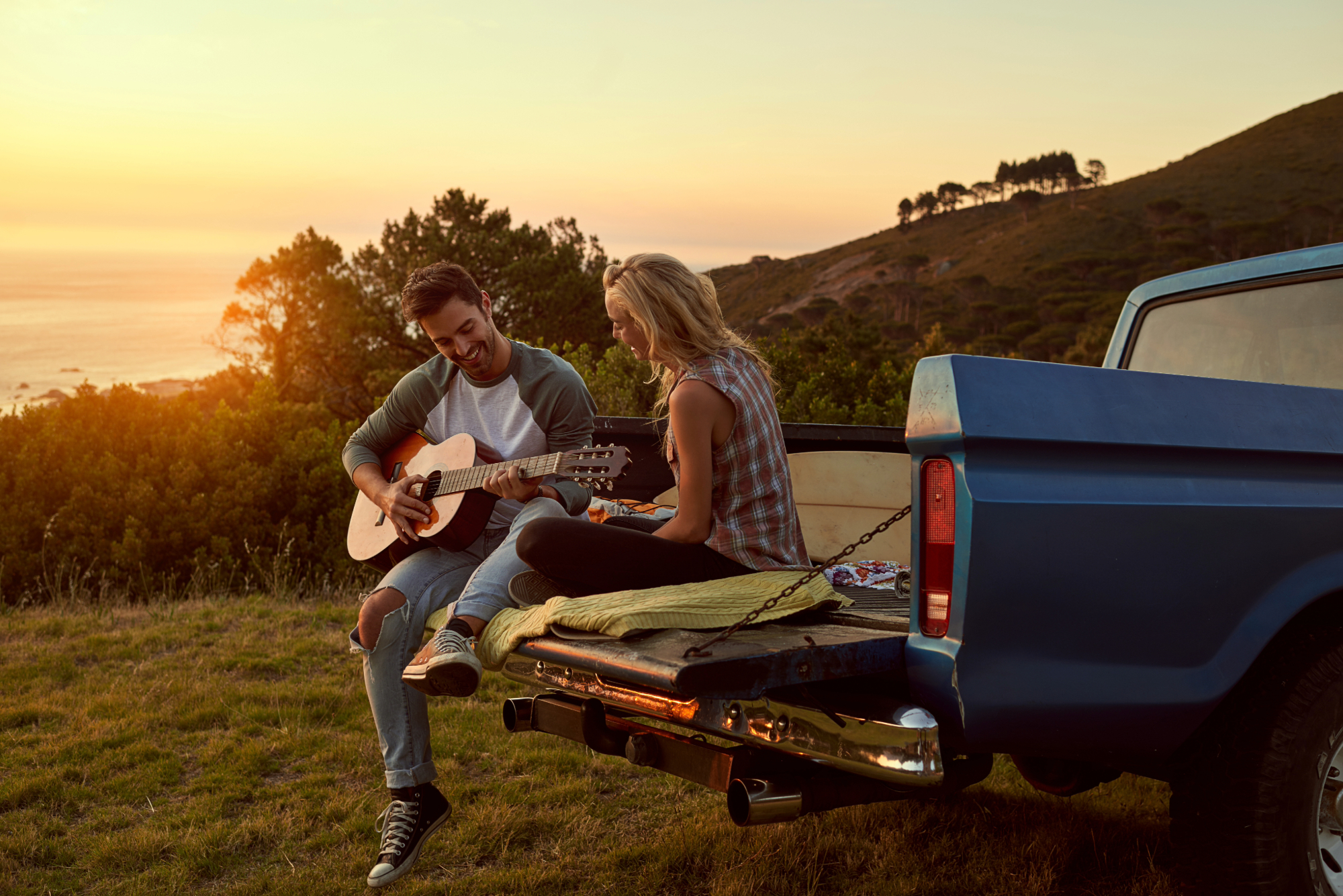 couple sitting on the tailgate of an old truck in front of a sunset while he plays guitar