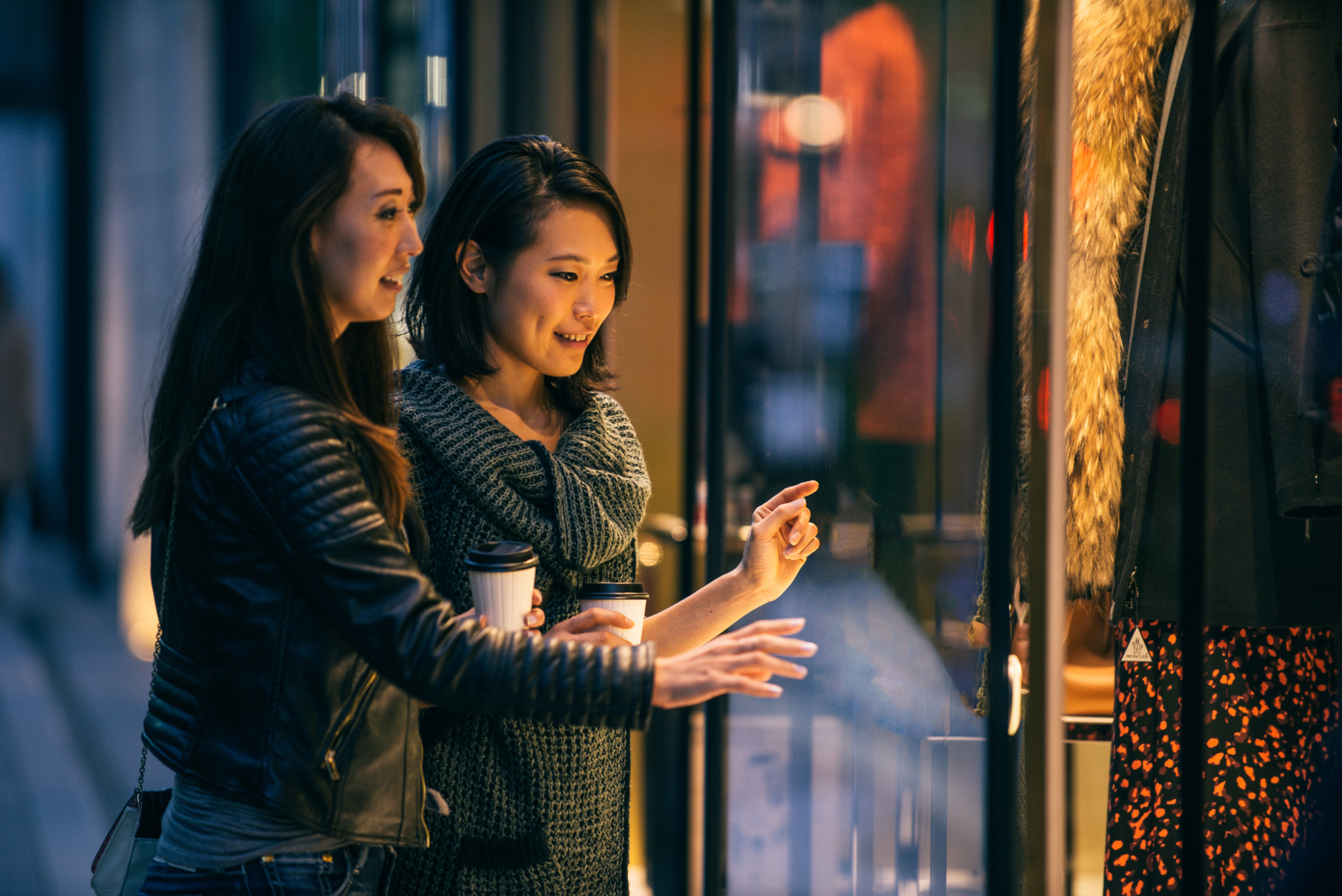 two women window shopping at night, pointing at something through the outdoor glass of a store