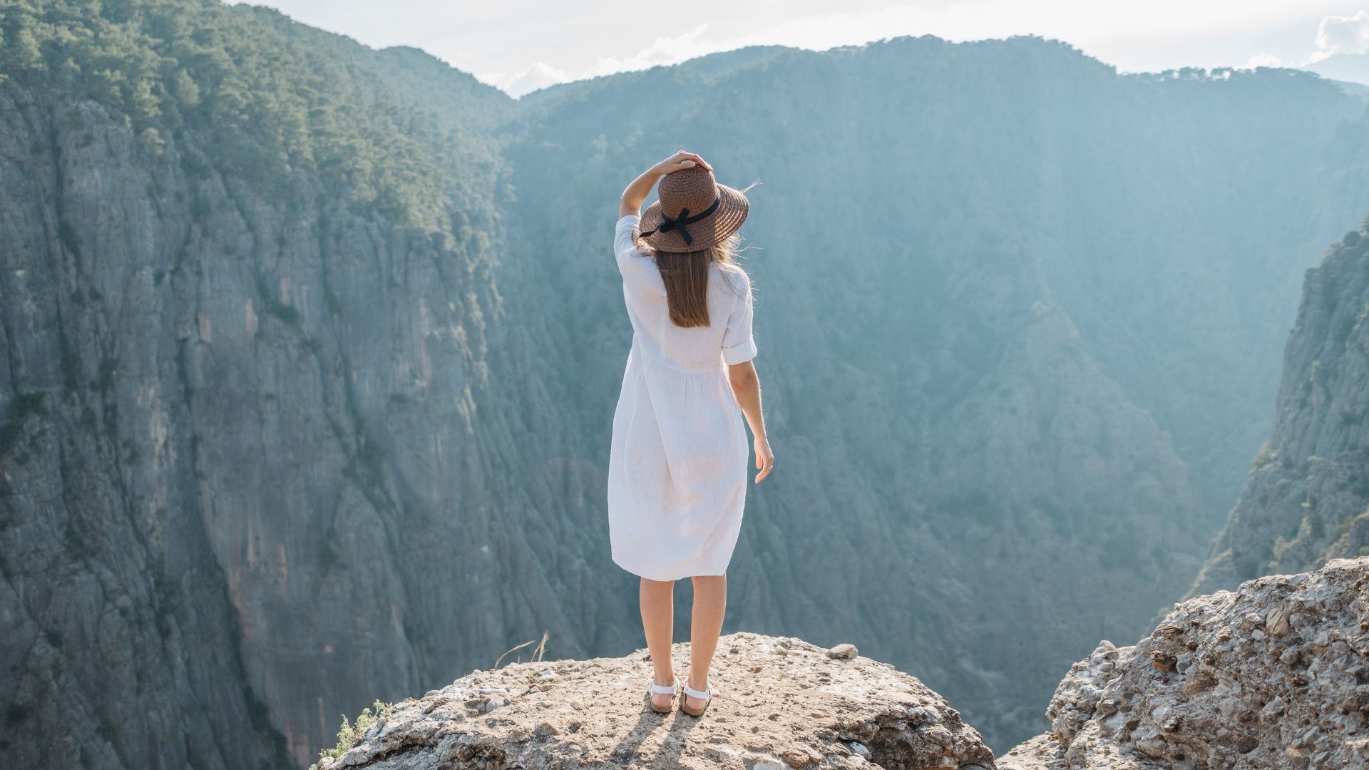woman in white dress and a tan sun hat overlooking a green canyon with gray rock walls during the daylight