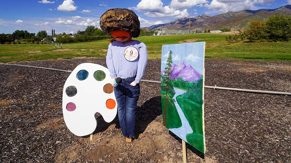 A Bob Ross themed scarecrow display with painter, palette, and painting.