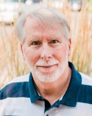 Photo of Tim Keady, male with white hair and trimmed beard, wearing a white and navy block striped shirt