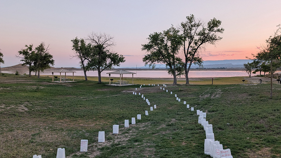 White luminaria bags line a curving path through a lakeside park at sunset.