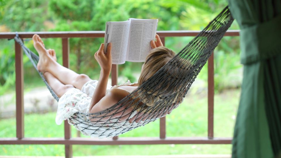 A women sitting in a hammock on a porch and reading a book.