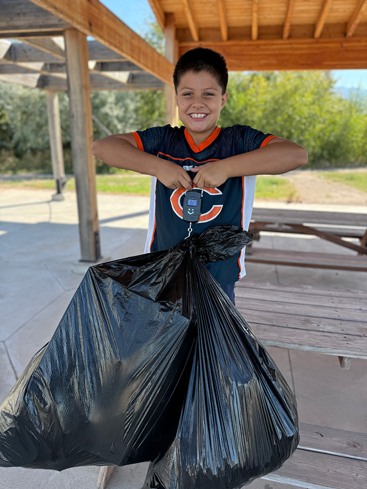 A smiling boy in a football jersey holds a trashbag on a scale for weighing