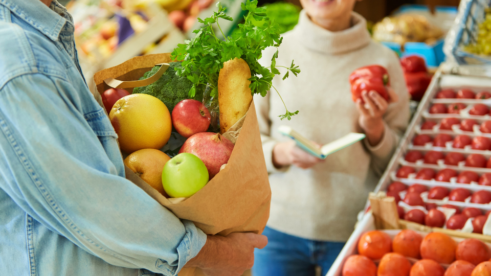 People in the produce section of a grocery store with a paper bag full of items.