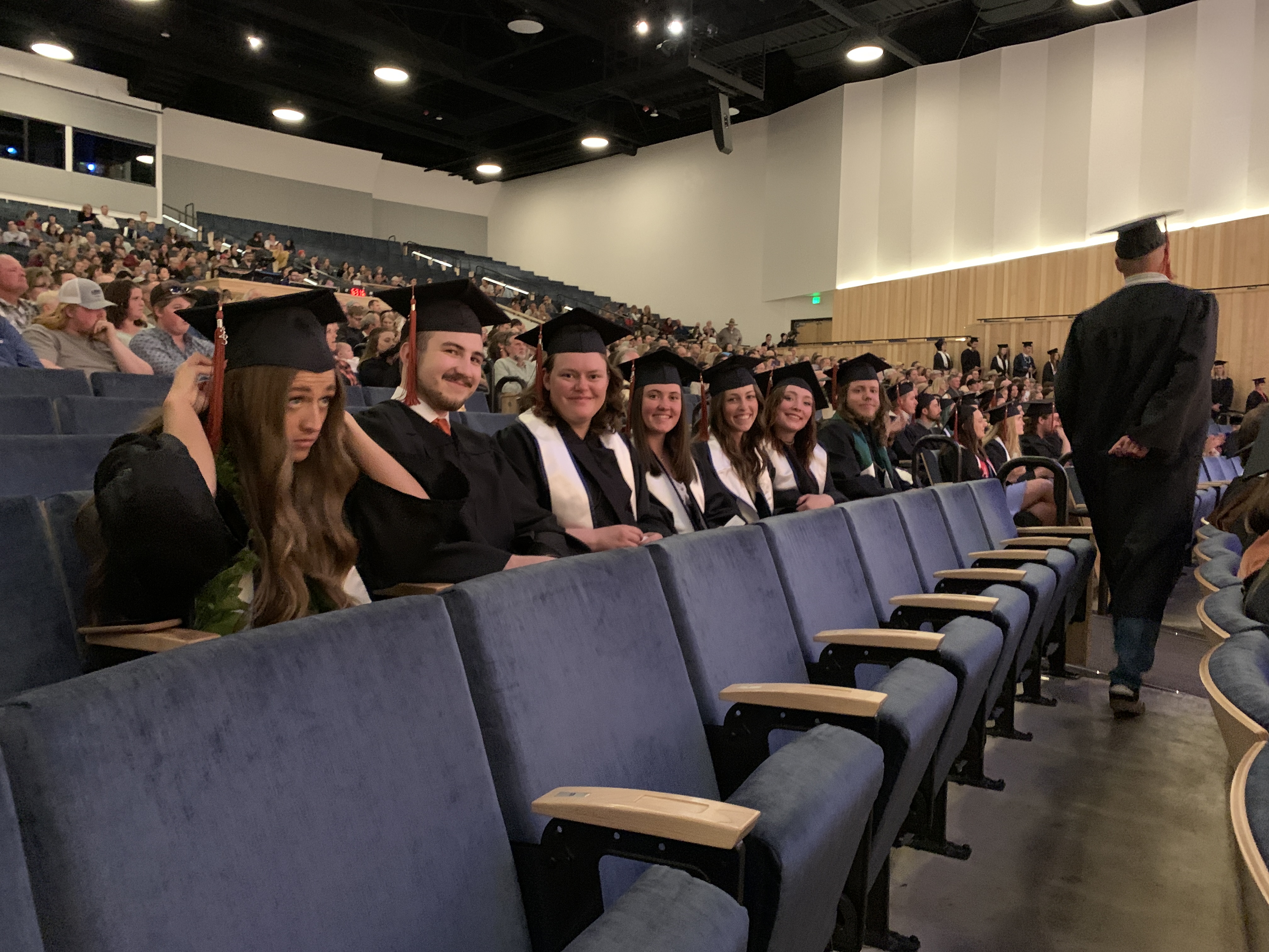 A row of students wearing graduation regalia sit in a row in an auditorium.