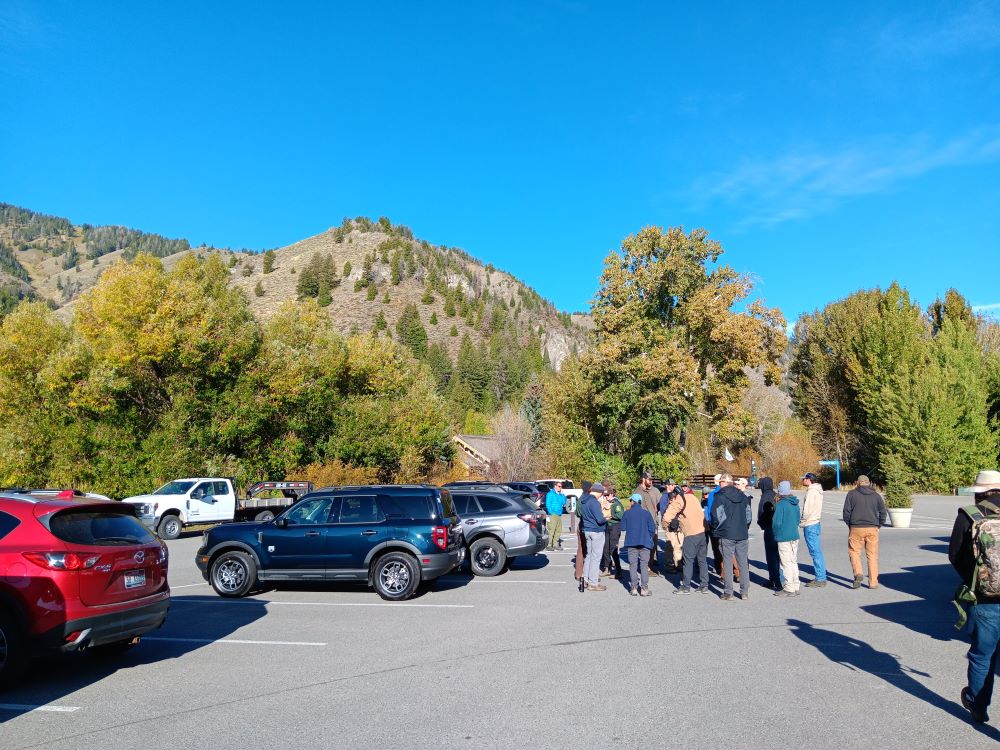 A group of people stand in a parking lot. There are trees and a mountain in the background.