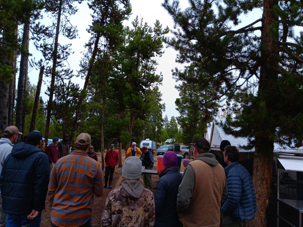 A group of people stand in a circle. There is a large white tent and several trucks in the background.