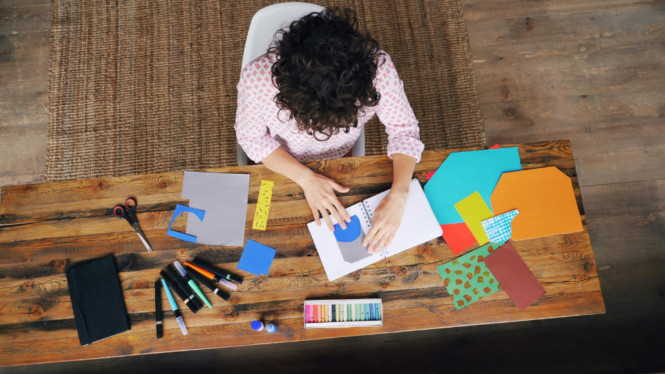 Person creating a paper craft project at a table covered with colorful paper, markers, scissors, and art supplies.