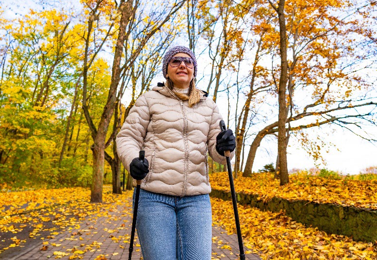 A women with walking sticks on the sidewalk with orange and yellow leaves falling from trees.