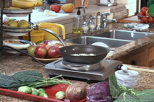 A kitchen counter with fresh vegetables, fruits, and a skillet on a portable stove, preparing a meal.