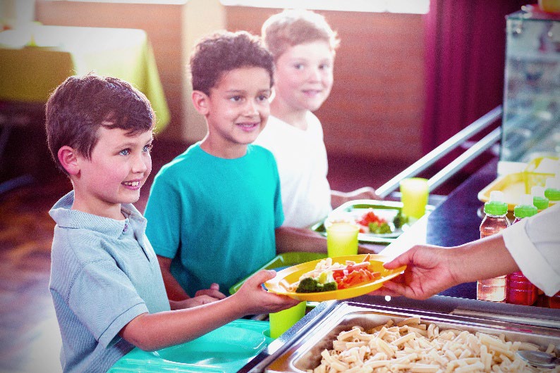 Three yound boys with trays in a lunch room being handed plates of vegitables
