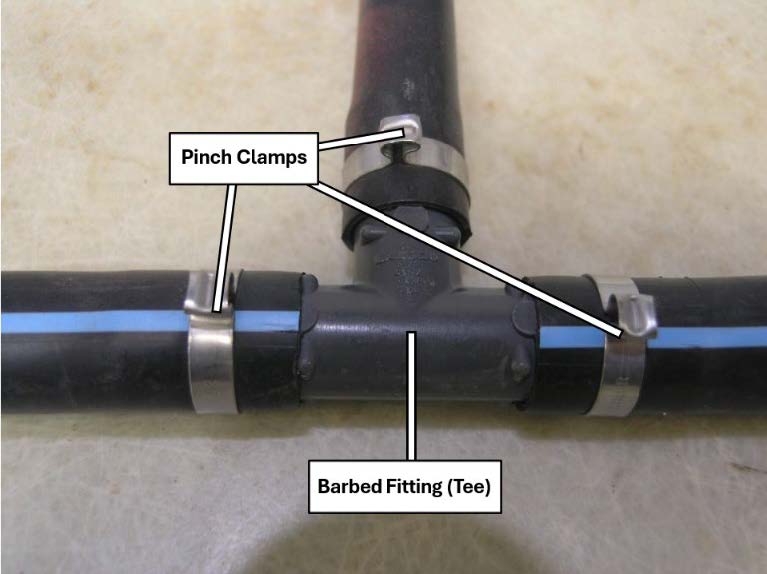 Close-up of a black poly irrigation tubing tee connection with three metal pinch clamps securing the tubing. Labels identify the pinch clamps and the barbed tee fitting that joins the three tubing sections.