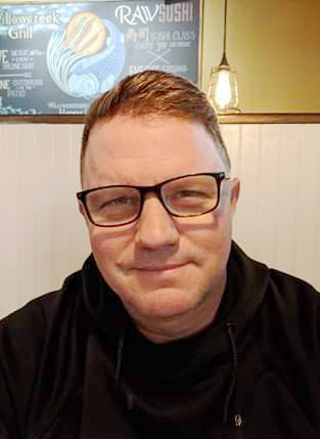 Robert Amis simling at the camera, wearing glasses and a black hoodie