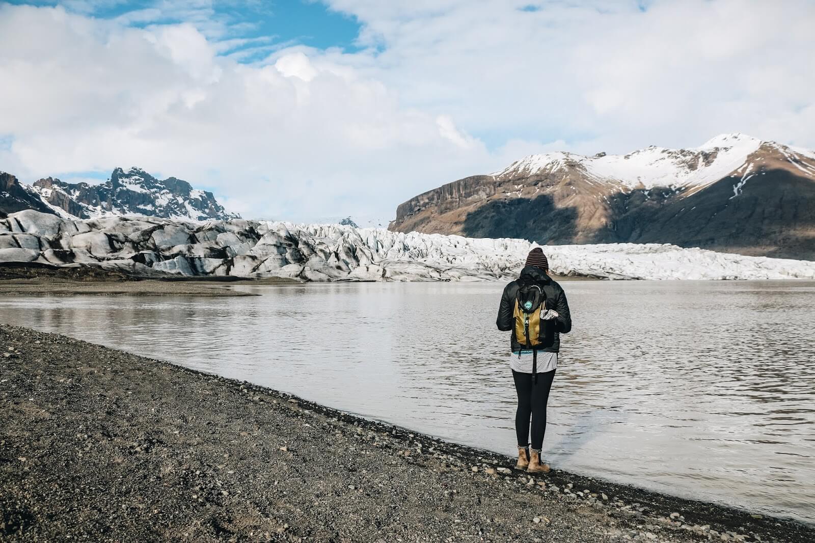 Terra standing in outdoor gear, facing a lake, glaciers and mountains.
