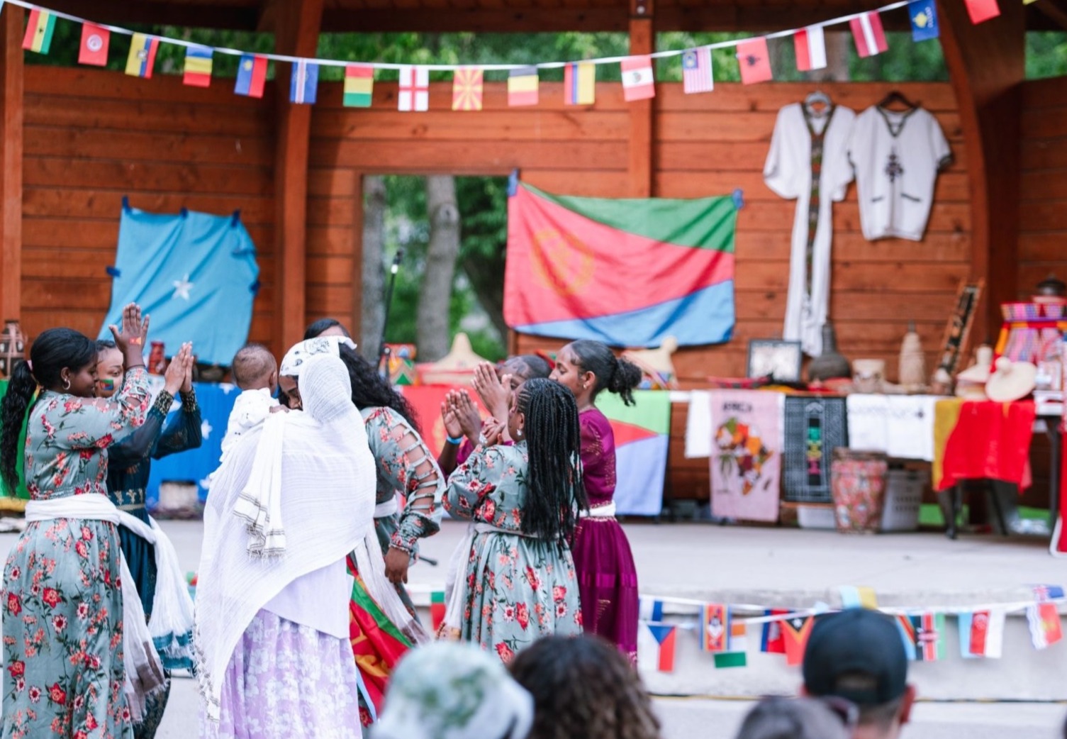 Group of women and girls in colorful traditional clothing performing a cultural dance on an outdoor stage decorated with international flags, while an audience watches.