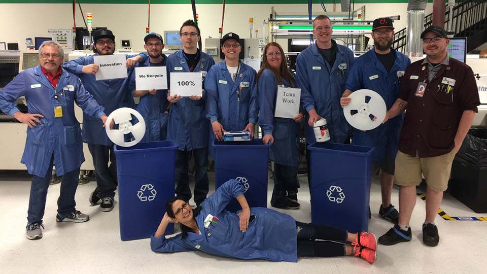 Employess in light blue work frocks posing around recycle bins holding recyclable items and holding signs that say, innovation, we recycle, 100% and team work