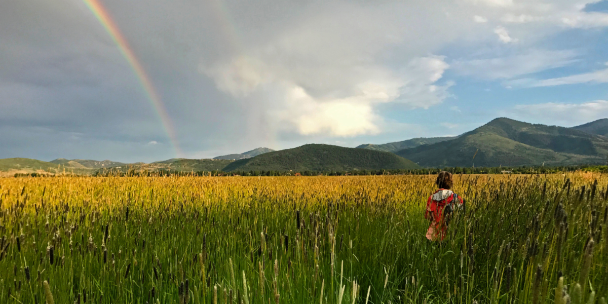 Child running through yellow grass towards mountains and rainbow