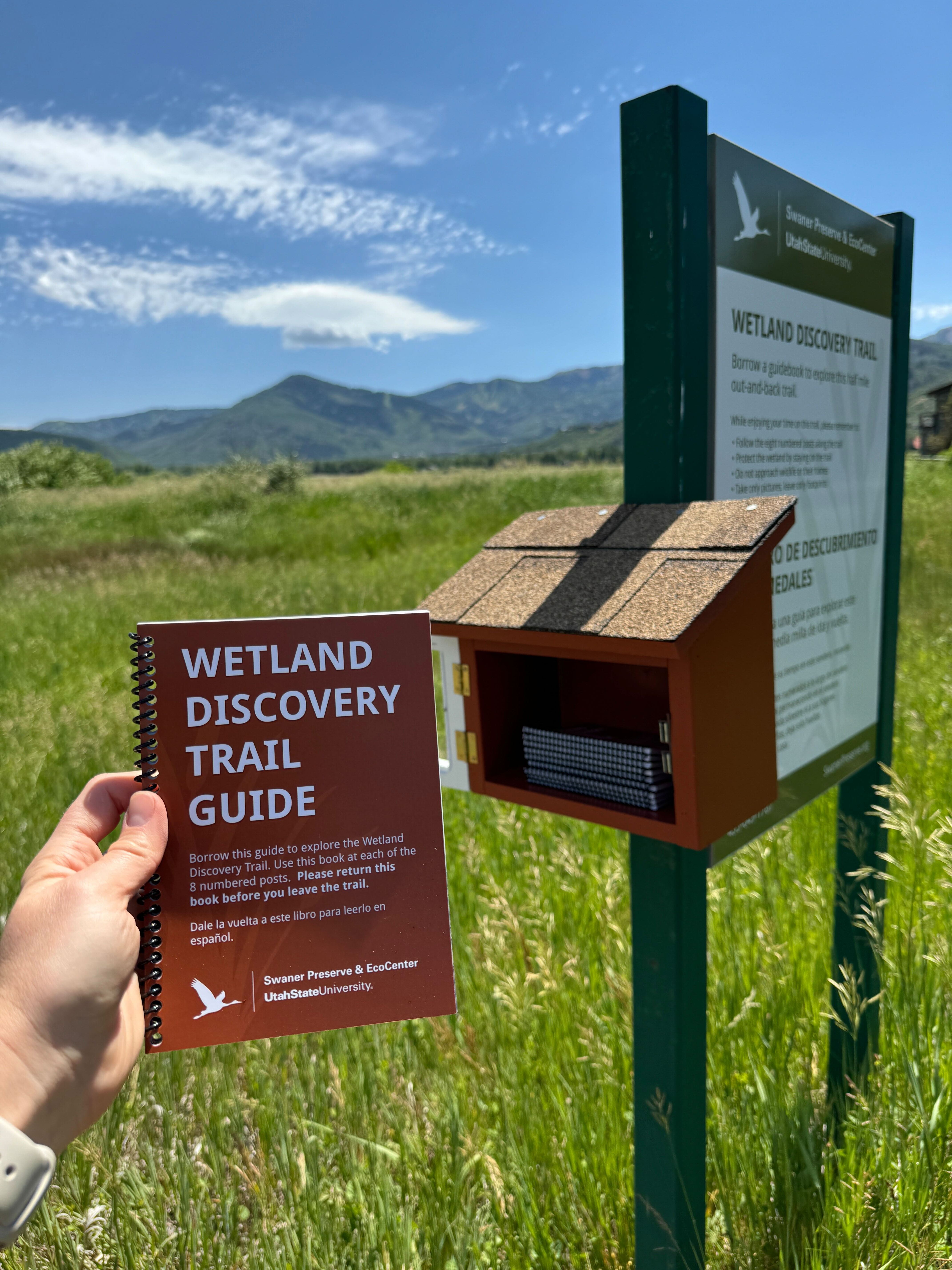 Someone holding an orange booklet titled “Wetland Discovery Trail Guide” in front of the trail sign and nature preserve and Wasatch mountains in the background 