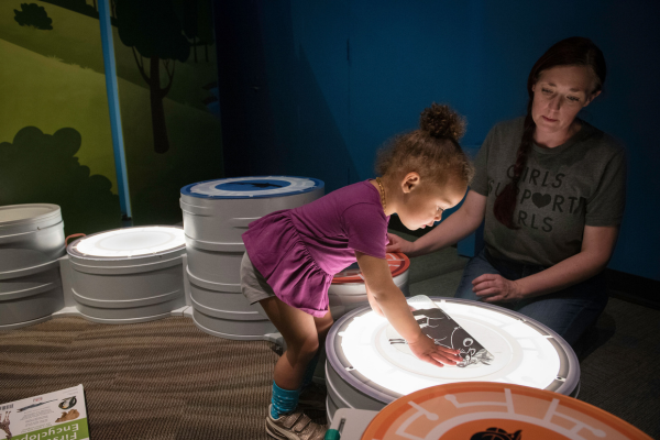 Child crouching down to put paper on a light-up small table like surface with the help of an adult.