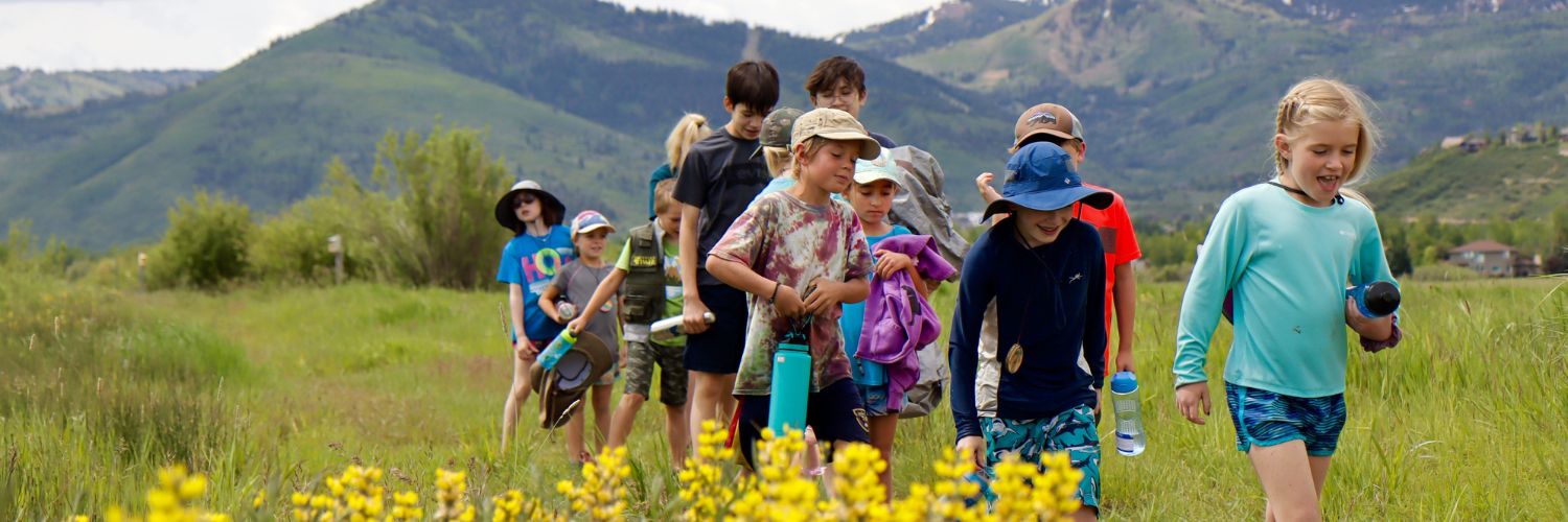 On a beautiful summer days, kids walking down flat grass path, laughing and smiling. Wasatch mountains are behind them and yellow rabbit brush is in front of them.